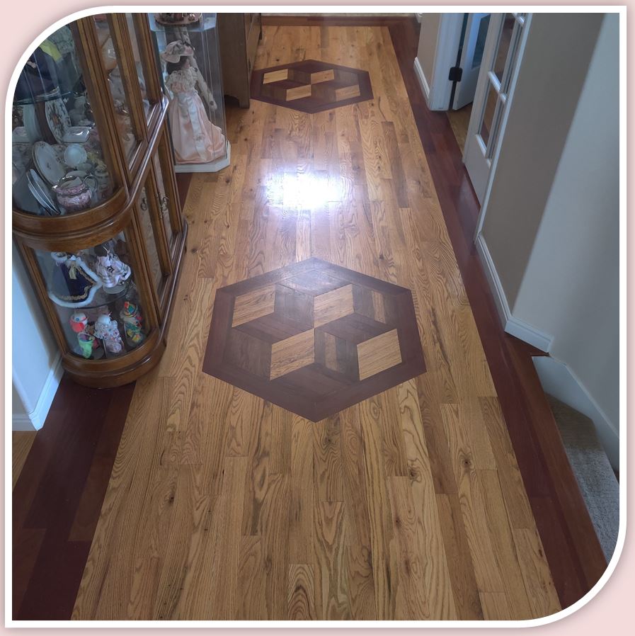 a solid red oak floor of custom design with Brazilian cherry border and inlay medallions made from Brazilian cherry, walnut and red oak, installed by Hardwood Floorist in a customer's entry hall. The hall is furnished with a curio cabinet, dolls and dishes. There is a large, elegantly dressed doll standing next to it.