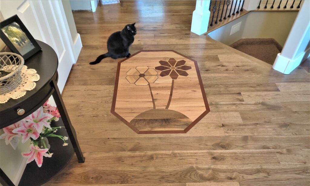 a hickory solid wood floor with a custom designed inlay medallion, picturing 2 flowers made from Santos mahogany, maple, walnut, and American cherry, on a red oak background, with a Brazilian cherry border, installed by Hardwood Floorist in a customer's entry way. The customer's beautiful black cat is sitting on the floor next to the medallion, looking down at it.