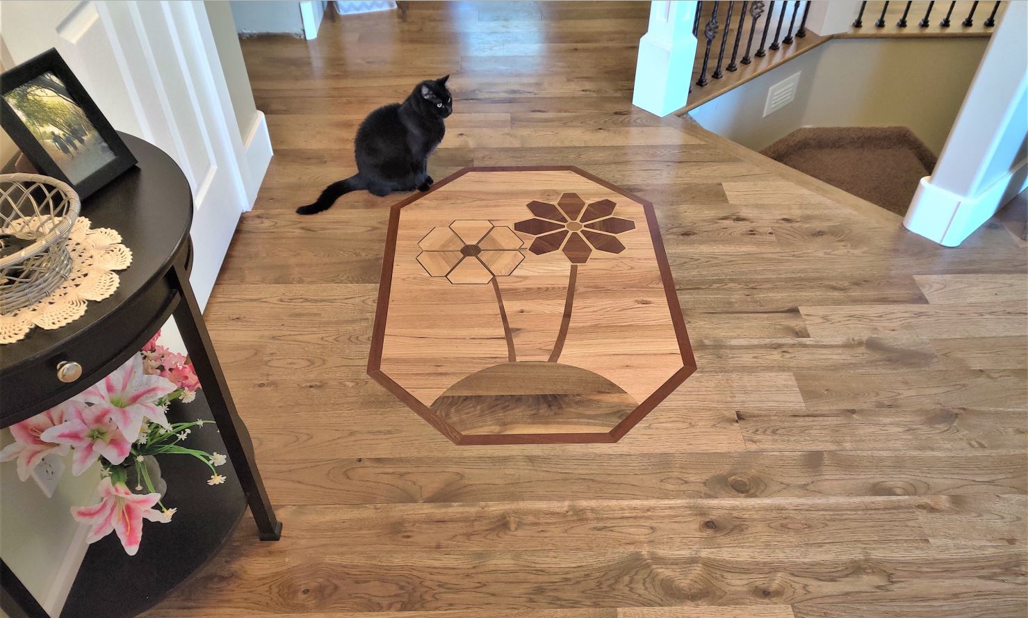 a hickory solid wood floor with a custom designed inlay medallion, picturing 2 flowers made from Santos mahogany, maple, walnut, and American cherry, on a red oak background, with a Brazilian cherry border, installed by Hardwood Floorist in a customer's entry way. The customer's beautiful black cat is sitting on the floor next to the medallion, looking down at it.