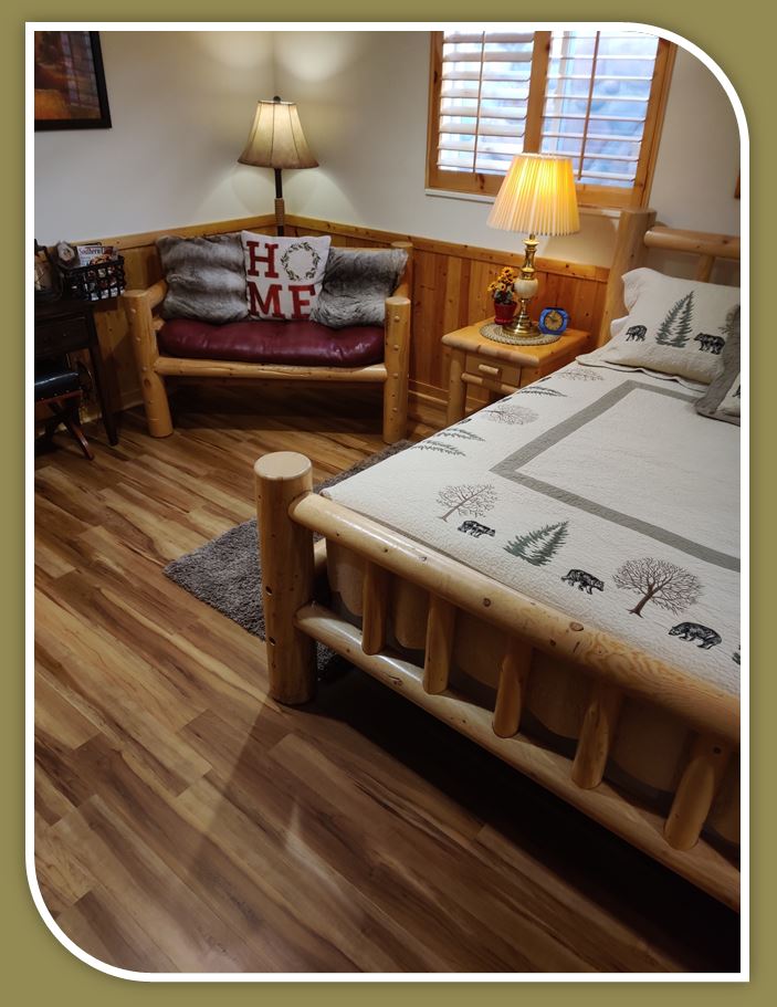 a waterproof, Red River hickory luxury vinyl plank (LVP) floor of traditional design, installed by hardwood floorist in a customer's guest bedroom. The bedroom is furnished with log cabin style bed, chair and lamps.