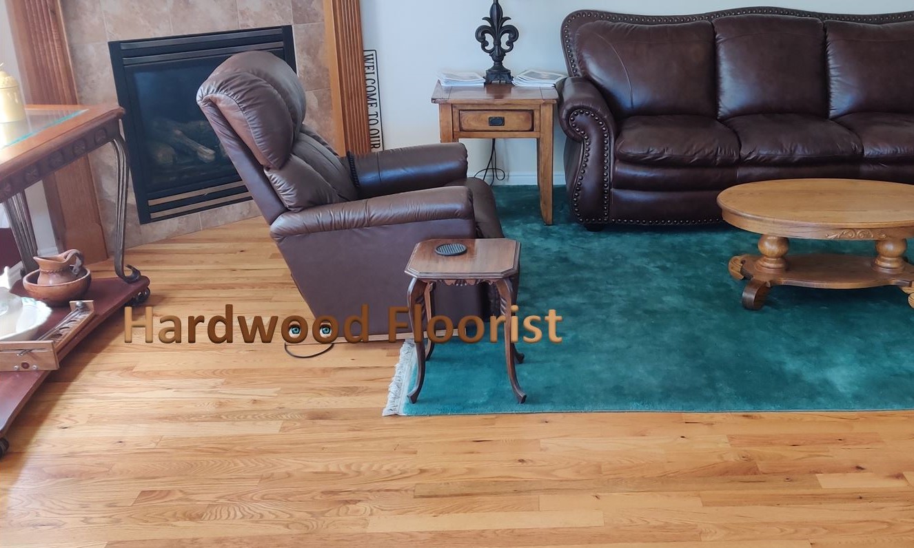 a red oak solid wood floor of traditional design, installed by Hardwood Floorist in a custommer's living room. The room has a fire place and is furnished with studded leather sofa and recliner chair, and coffee and end tables, sitting atop a large aquamarine rug.