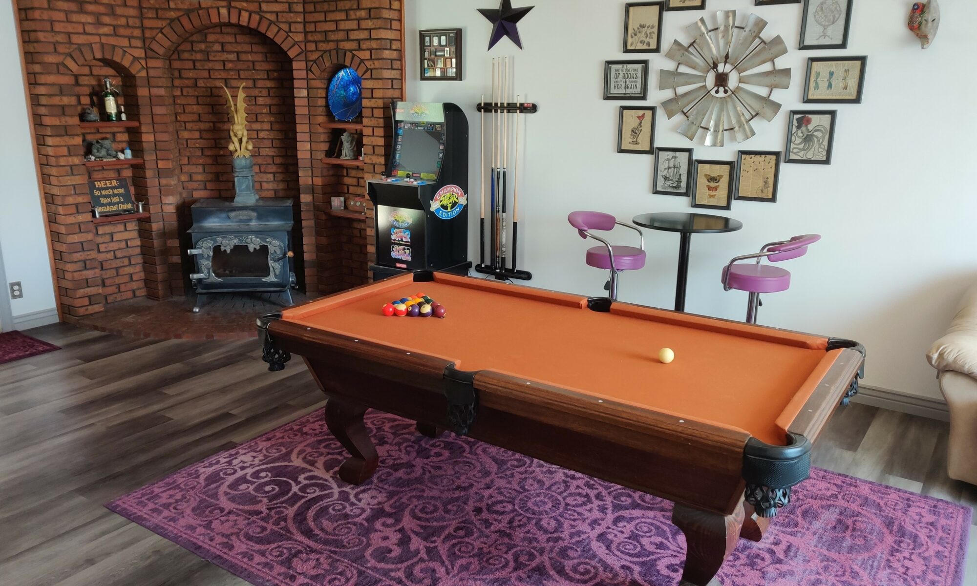 a stormy gray oak, waterproof, luxury vinyl plank (LVP) floor, installed by Hardwood Floorist in a customer's game room. The room has a large brick mantle with a wood burning stove, and contains a billiards table and a small, round, tall table and chairs set. There is interesting art on the walls, including a sign that says: "Beer: so much more than just a breakfast drink".